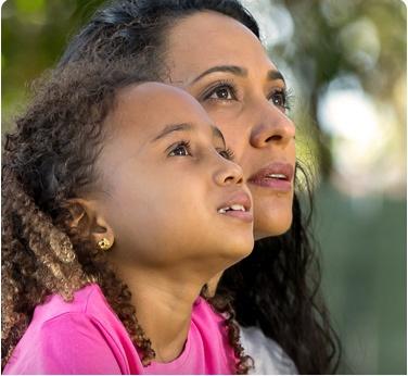 Mother and daughter smiling together outdoors, enjoying a sunny day with a scenic background of greenery and blue sky.